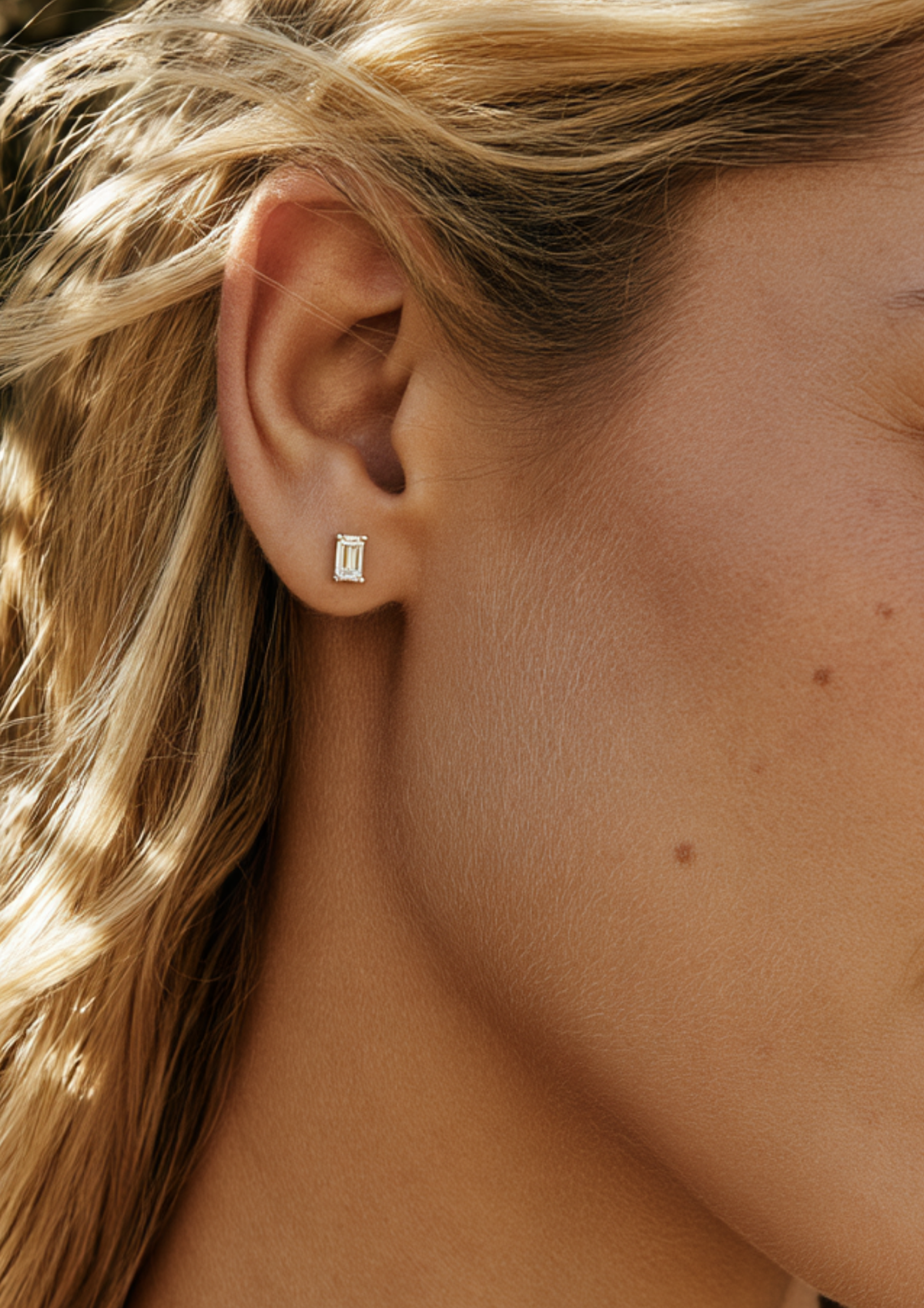 A close-up of a model's ear wearing a small rectangular silver stud earring with a clear gemstone.