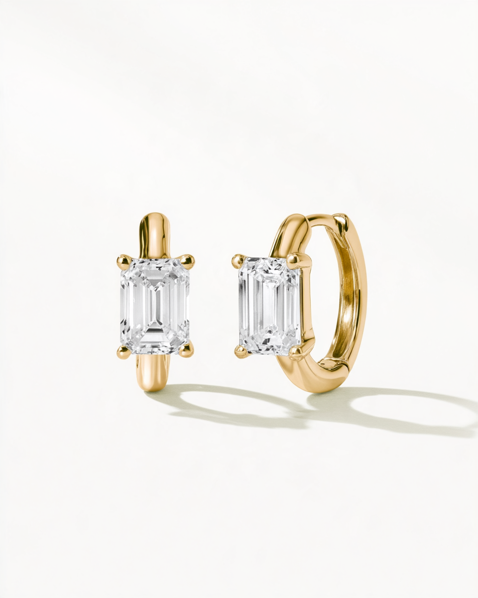A pair of gold huggie hoop earrings with rectangular emerald-cut clear gemstones, displayed on a clean white background with soft shadows.