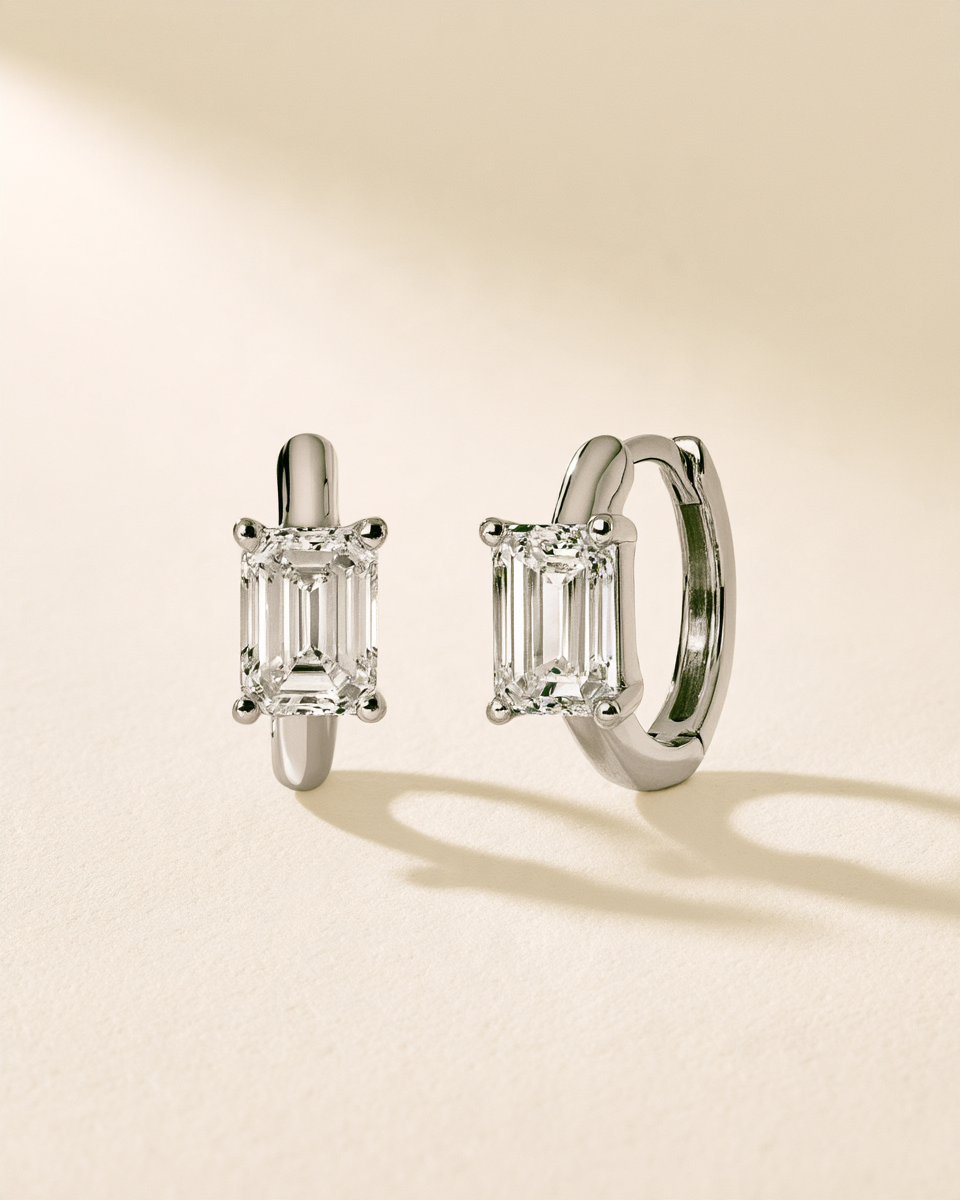 A pair of silver huggie hoop earrings with baguette-cut clear gemstones, displayed on a warm beige background with soft shadows.