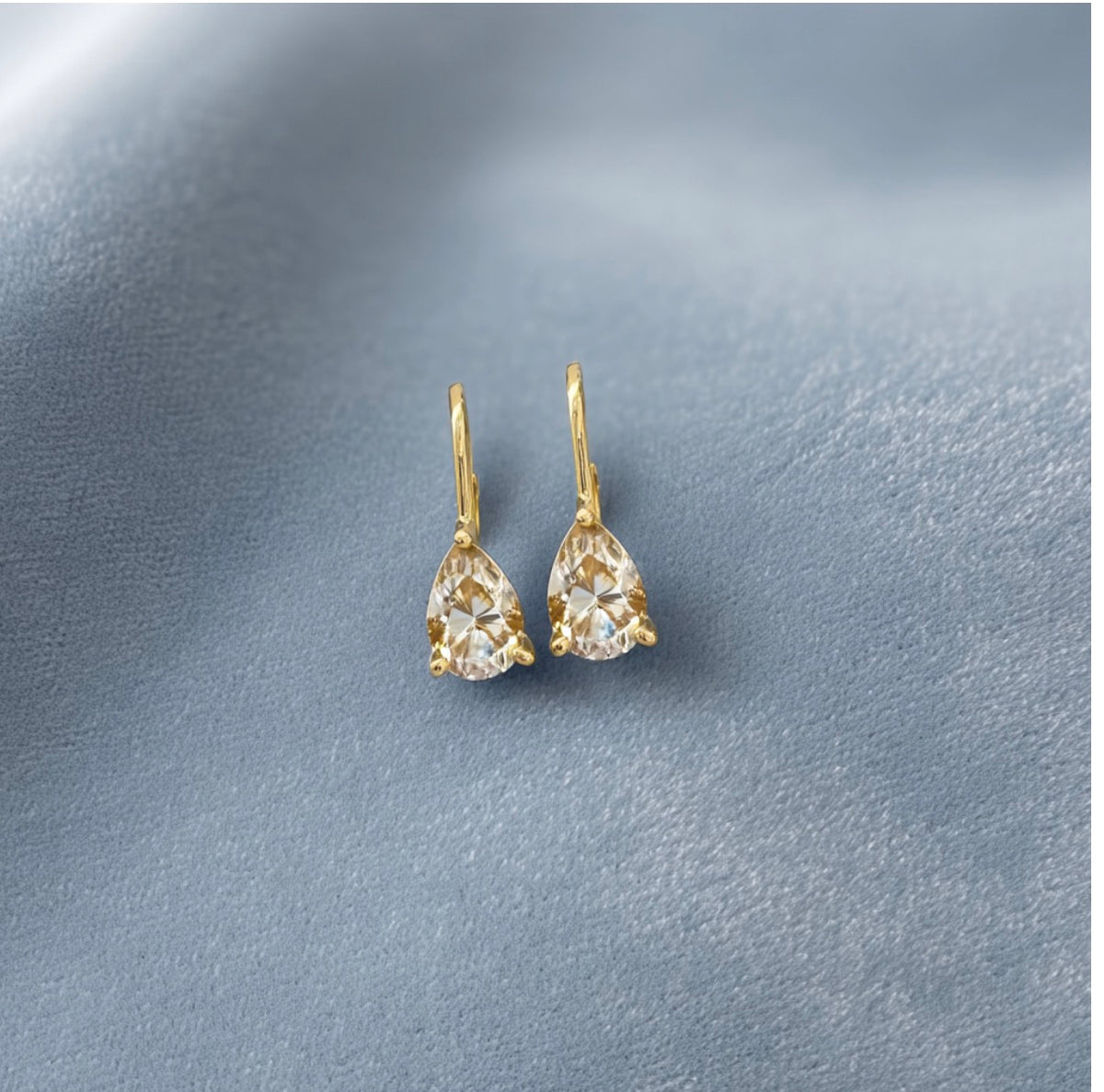 Violet Pear Earrings with champagne stones and gold-tone lever backs on a blue-grey surface.
